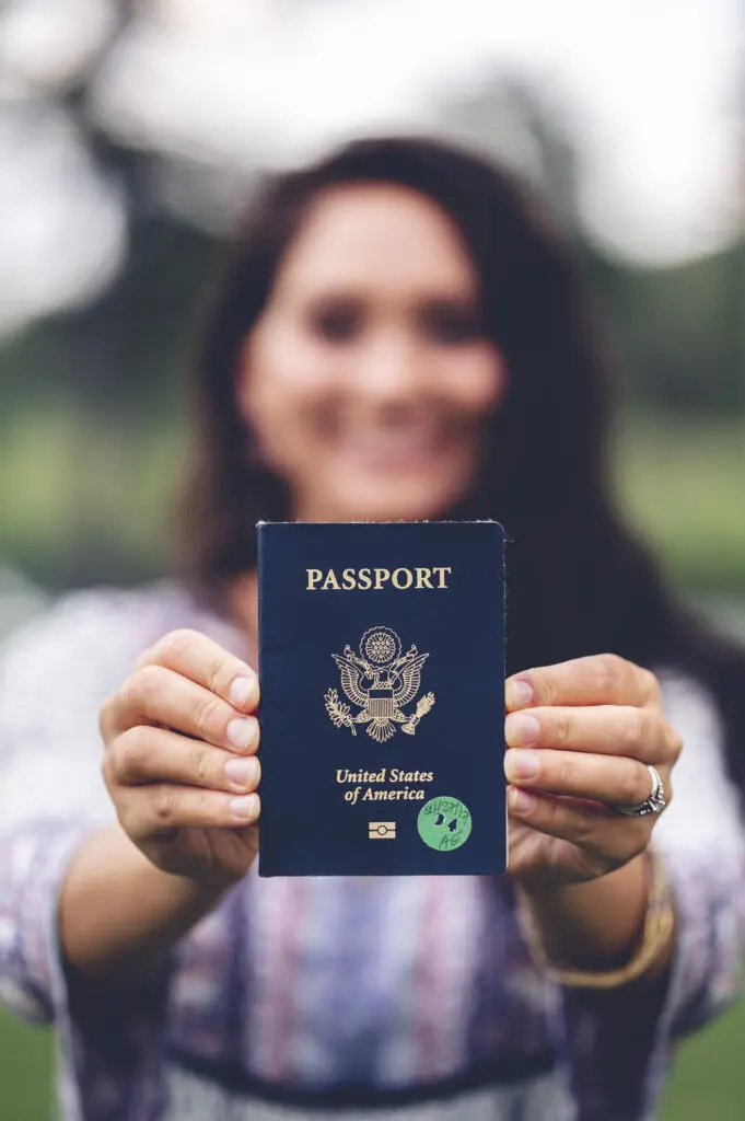 A person holds out a United States passport towards the camera, with the cover and seal clearly visible—perhaps preparing to apply for a Tourist Visa.