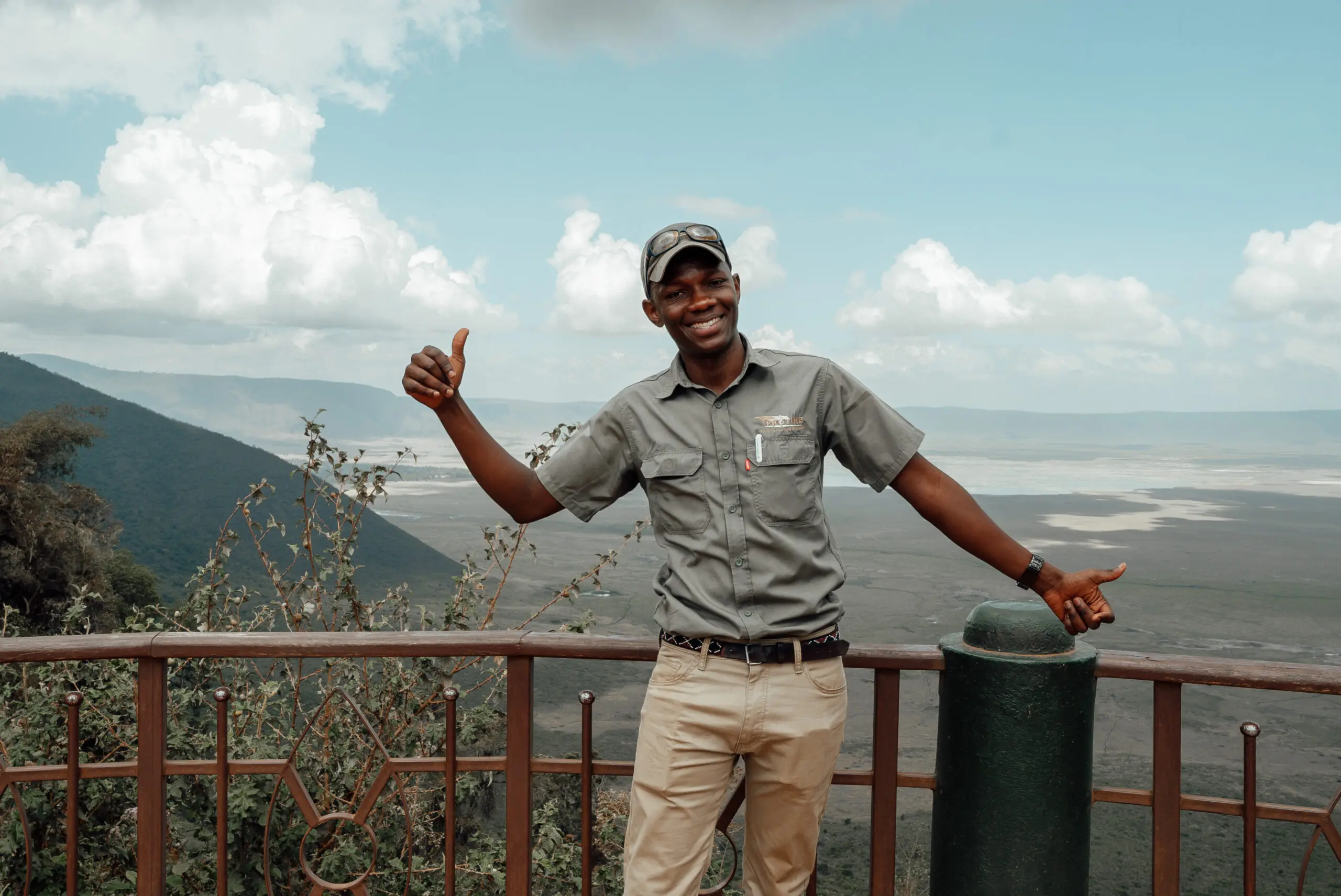 A person in a cap and casual clothing stands by a railing, smiling and giving a thumbs-up, with a vast landscape and cloudy sky in the background—a perfect moment to capture before you book your trip.
