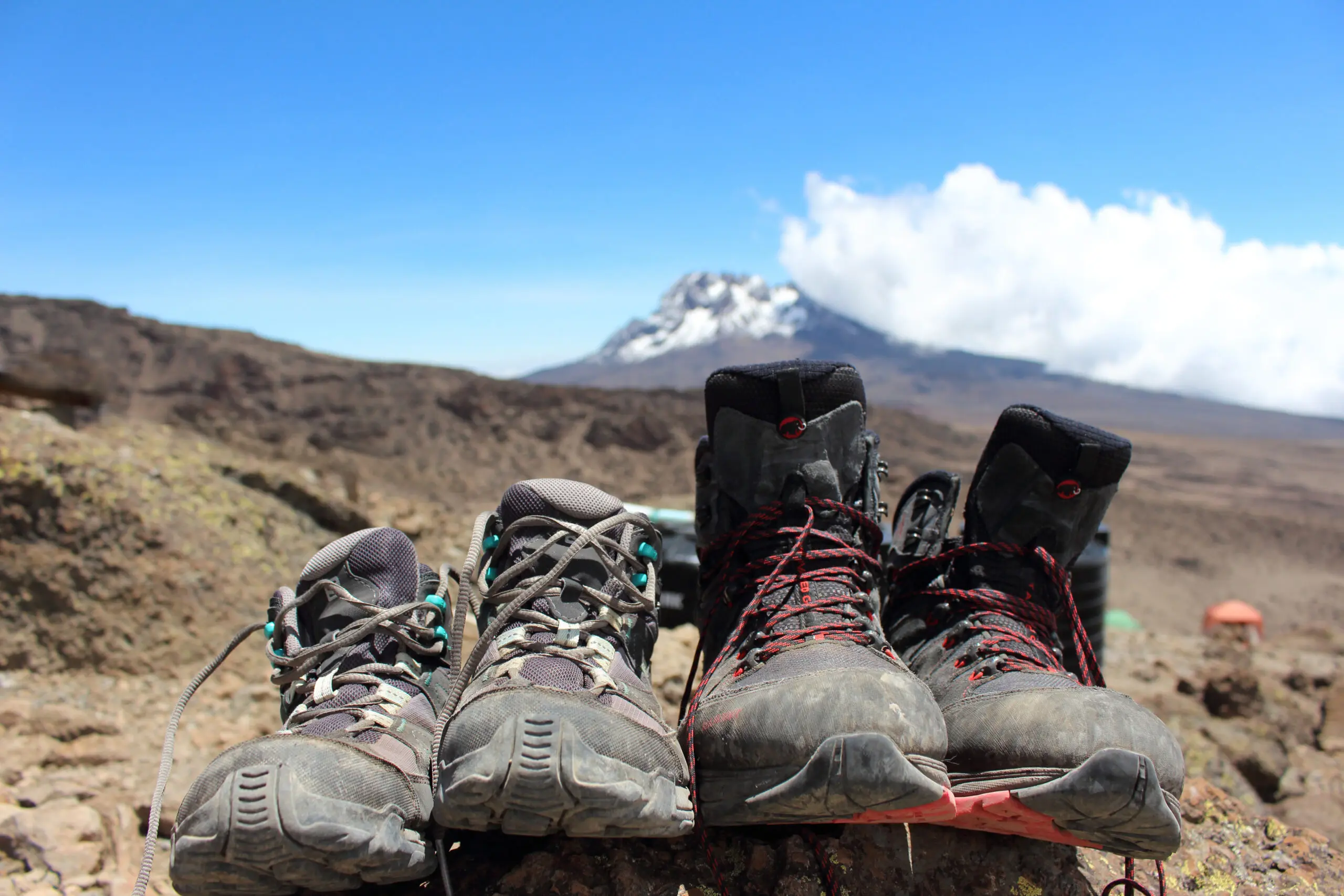 Two pairs of hiking boots rest on rocky ground, framed by a mountain peak and blue sky—capturing the spirit to hike Mount Kilimanjaro.