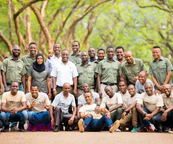 A group of people in casual attire, maintaining social distancing, pose outdoors on a path lined with trees, mindful of COVID-19 precautions.