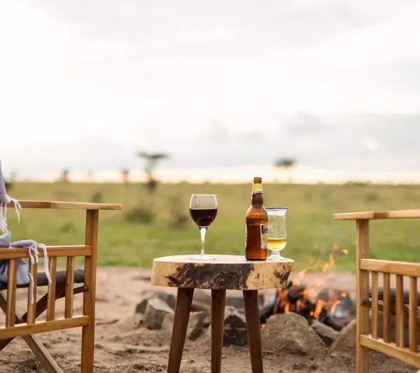 Two wooden chairs face a small table with drinks beside a campfire, set in an open grassy landscape, providing the perfect spot to unwind and travel safely.