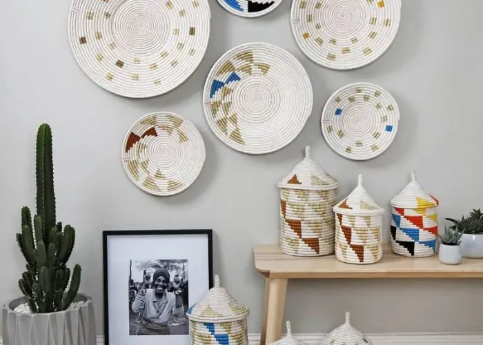 Woven baskets with geometric patterns hang on a wall, embodying a perfect touch for any Holiday Gift Guide. Below, covered baskets and a framed photo sit on a bench, next to a potted cactus.