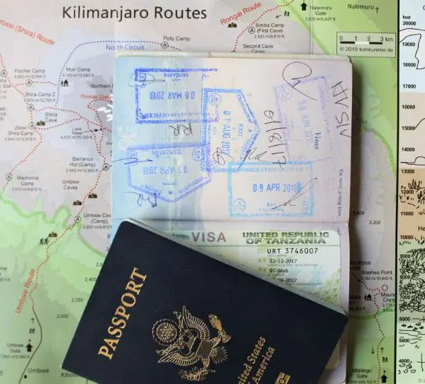 A U.S. passport with a Tanzanian visa and proof of a recent COVID-19 test lies open on a map, detailing Kilimanjaro routes and climate zones.