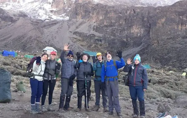 A group of seven people in hiking gear pose and wave at a mountainous campsite, reminiscent of Kilimanjaro, with tents and rocky terrain in the background.