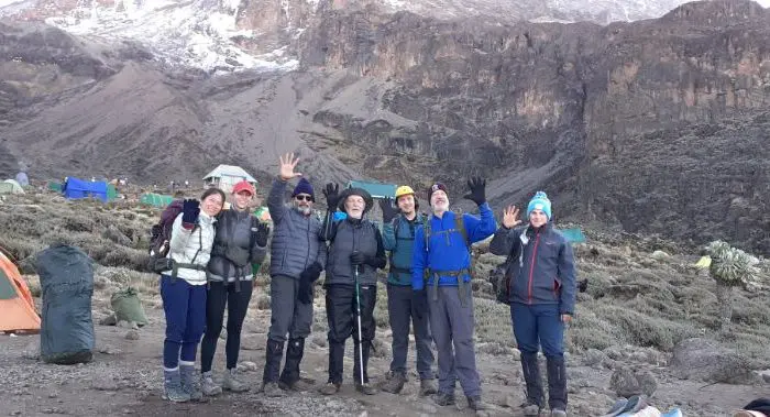 A group of seven people in hiking gear pose and wave at a mountainous campsite, reminiscent of Kilimanjaro, with tents and rocky terrain in the background.