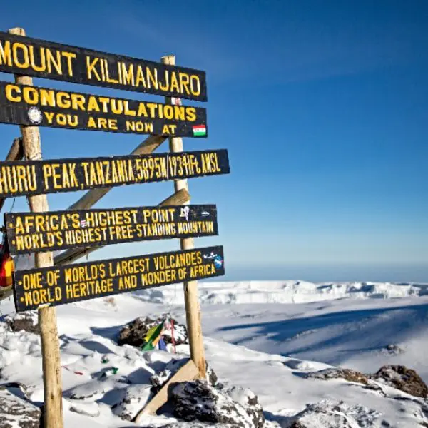 A wooden sign on the snowy slopes of Mount Kilimanjaro marks Uhuru Peak at 5895m, the highest point in Africa. The breathtaking view includes a snow-covered landscape and a clear blue sky, reminiscent of the vast beauty seen across the Serengeti.