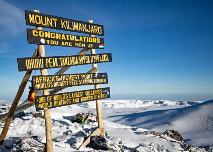 A wooden sign on the snowy slopes of Mount Kilimanjaro marks Uhuru Peak at 5895m, the highest point in Africa. The breathtaking view includes a snow-covered landscape and a clear blue sky, reminiscent of the vast beauty seen across the Serengeti.