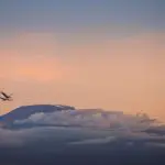 An airplane glides gracefully past a mountain peak, silhouetted against a cloudy sky at sunset, evoking dreams of traveling to Tanzania's breathtaking landscapes.