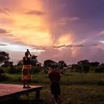 Two people stand on a wooden platform in a national park, photographing a vibrant sunset over the lush landscape with scattered trees.