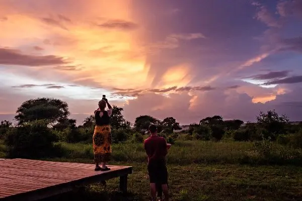 Two people stand on a wooden platform in a national park, photographing a vibrant sunset over the lush landscape with scattered trees.