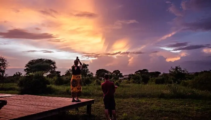 Two people stand on a wooden platform in a national park, photographing a vibrant sunset over the lush landscape with scattered trees.