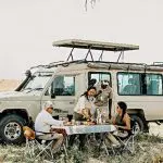 A group enjoys a picnic beside a safari vehicle in the grassy expanse of a national park's savanna.