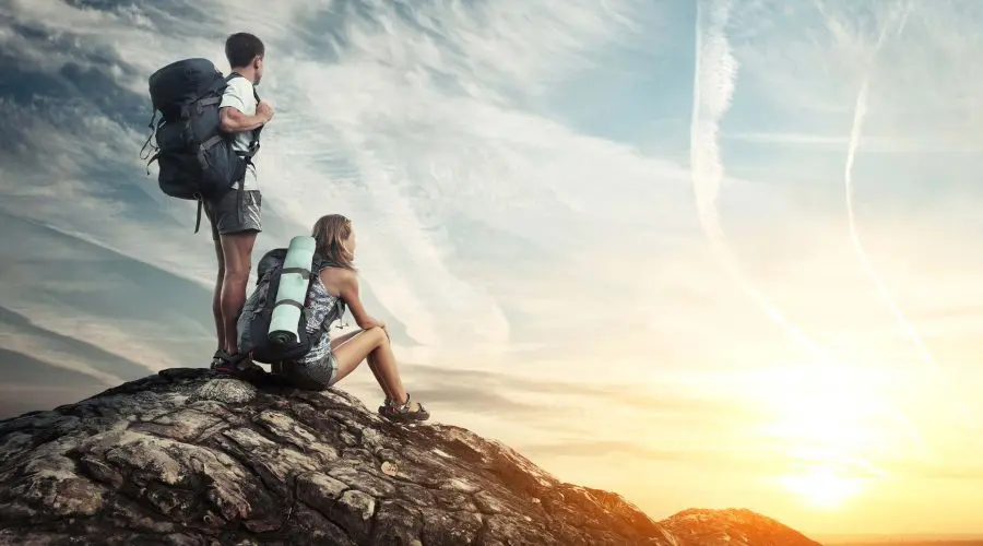 Two hikers with backpacks stand and sit on a rocky hilltop, watching a sunrise with a sky streaked by clouds.