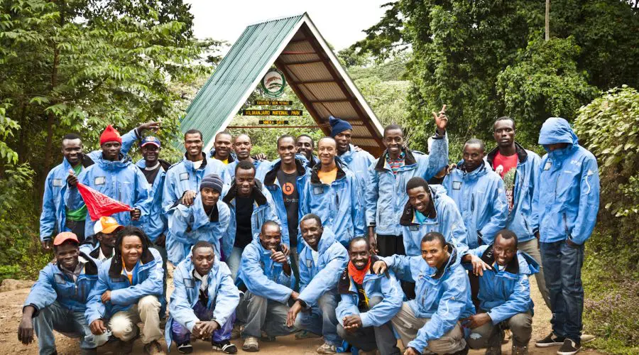 A group of people in blue jackets pose together outdoors near a triangular wooden structure surrounded by greenery.