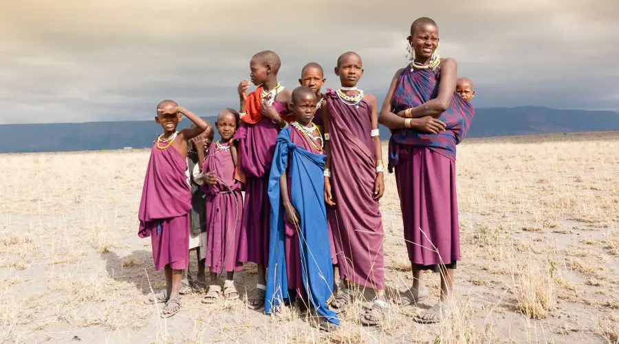 A group of people wearing traditional attire stand together on a dry, grassy plain under a cloudy sky.