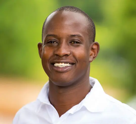 Smiling man in a white shirt with "Pristine Adventures and Safaris" logo stands against a blurred green background.