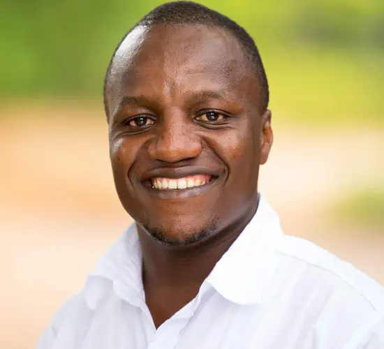 Smiling person in a white shirt with "Pristine Adventures and Safaris" logo, standing outdoors with a blurred green and beige background.