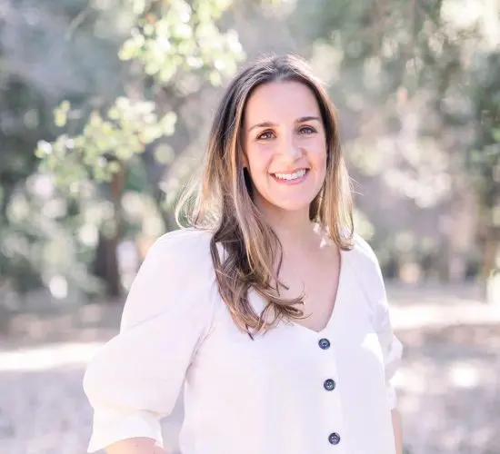 Woman smiling in a sunlit outdoor setting, wearing a white blouse and jeans with hands in pockets. Trees and greenery in the blurred background.