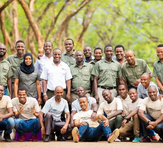 A diverse group of people, wearing uniforms and casual outfits, pose together smiling outdoors with a background of trees.