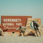 Three people pose energetically in front of the Serengeti National Park entrance sign on a sunny day.