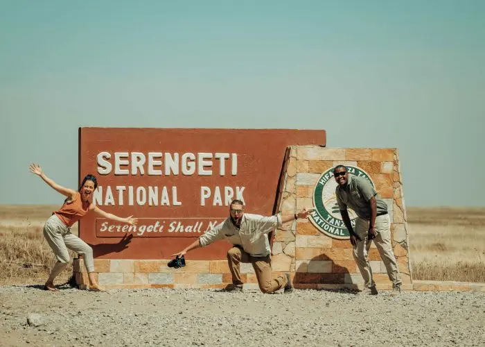 Three people pose energetically in front of the Serengeti National Park entrance sign on a sunny day.