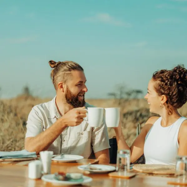 Two people sit at an outdoor table in a field, holding white mugs and smiling at each other, their laughter as warm as the Tanzanian sun. The table is set with plates and glasses, capturing the essence of this perfect stop on their travel itinerary.