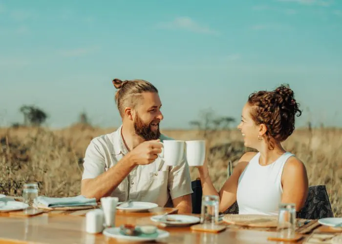 Two people sit at an outdoor table in a field, holding white mugs and smiling at each other, their laughter as warm as the Tanzanian sun. The table is set with plates and glasses, capturing the essence of this perfect stop on their travel itinerary.