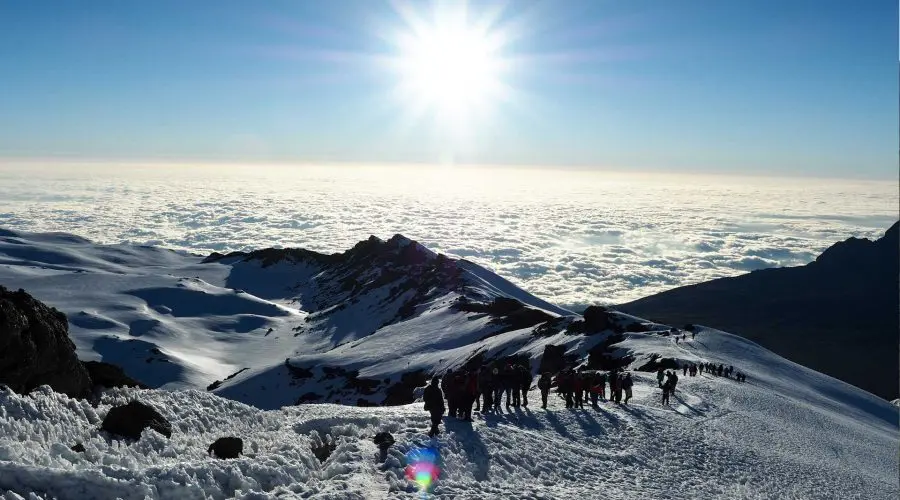 A group of climbers ascends a snowy mountain ridge under a bright sun, with a sea of clouds below.