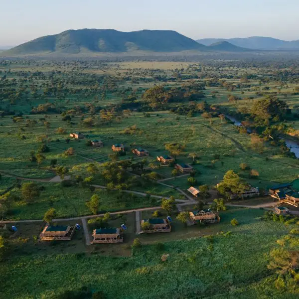 Aerial view of a lush green savanna landscape with a small lodge complex, surrounded by scattered trees and mountains in the background.