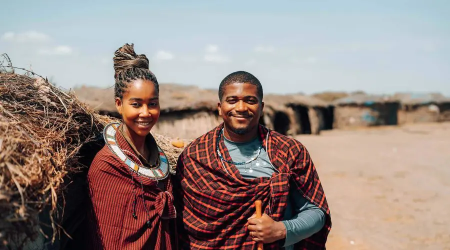 Two people wearing traditional Maasai attire stand outdoors, smiling. In the background are huts and a clear sky.