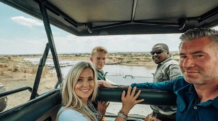 Four people in a safari vehicle, parked near a river in a savannah landscape. One woman and three men are smiling at the camera.