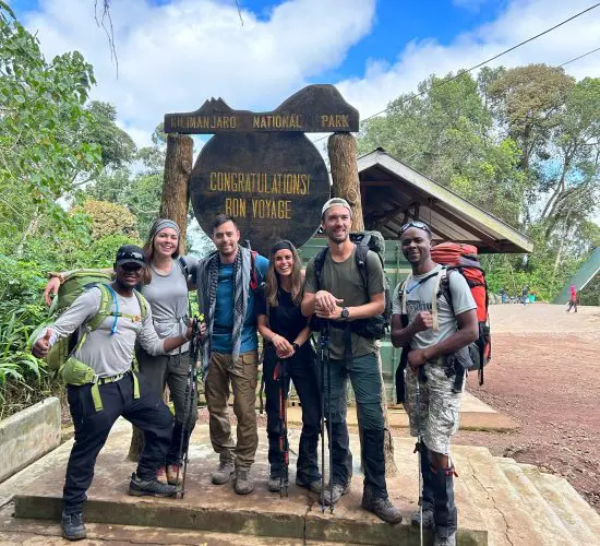 A group of hikers poses in front of the Kilimanjaro National Park sign, holding trekking poles and wearing outdoor gear.