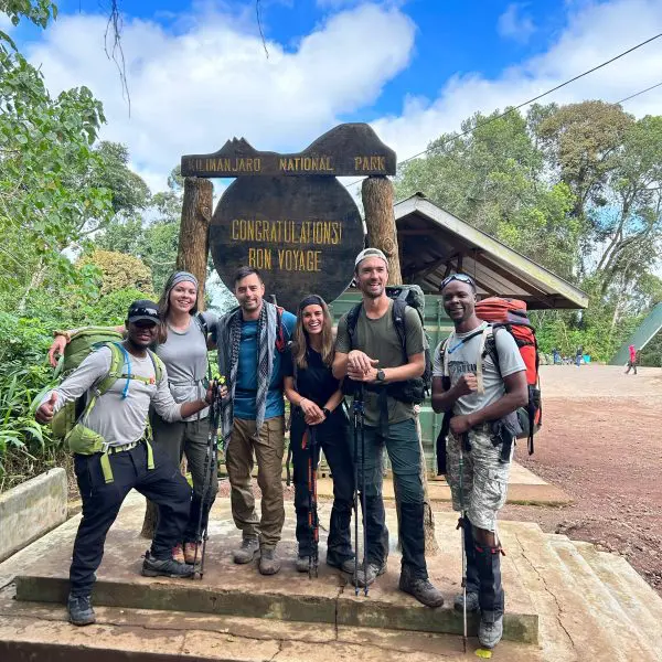 A group of hikers poses in front of the Kilimanjaro National Park sign, holding trekking poles and wearing outdoor gear.