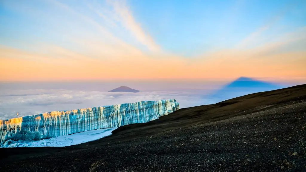 Ice cliff on a mountain slope with a view of clouds and two peaks in the background under a colorful sky at sunset.