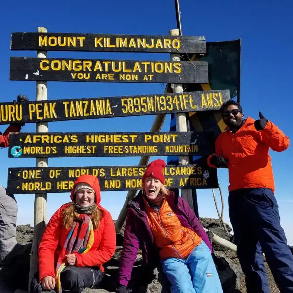 Four people in hiking gear stand in front of the Mount Kilimanjaro summit sign, celebrating their achievement on a clear day.