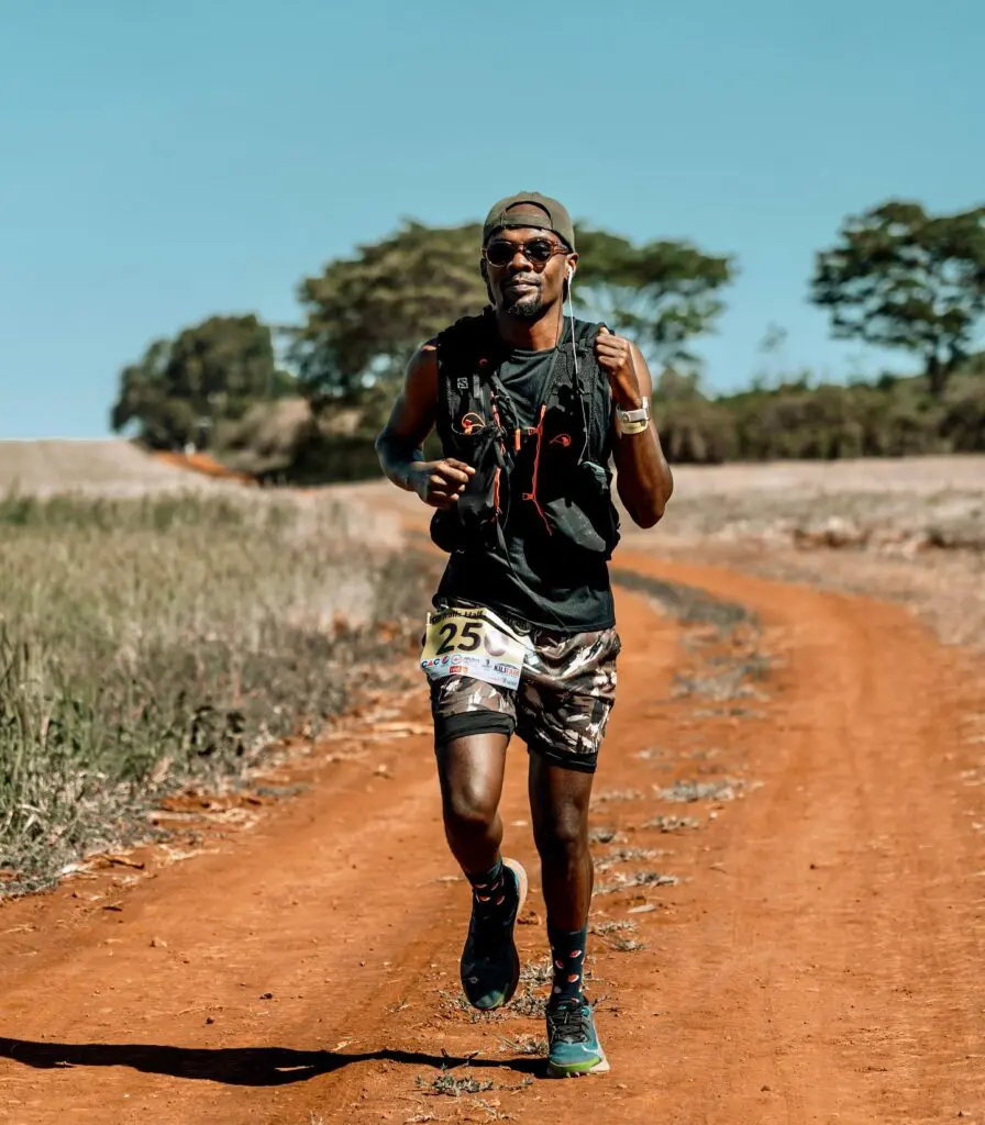 A person runs on a dirt path through a rural landscape under a clear sky, wearing athletic gear and a numbered race bib.