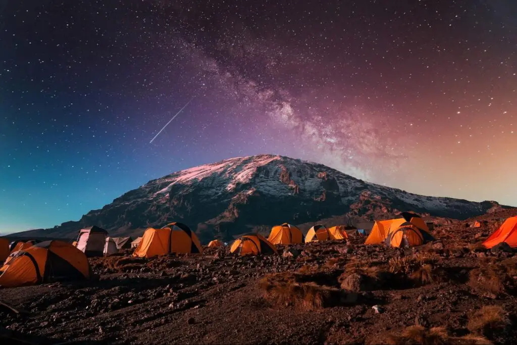 Orange tents set up on rocky terrain under a starry night sky with the Milky Way and a snowy mountain in the background.