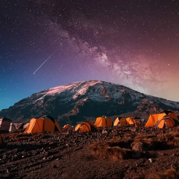 Orange tents set up on rocky terrain under a starry night sky with the Milky Way and a snowy mountain in the background.