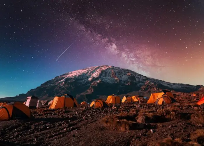 Orange tents set up on rocky terrain under a starry night sky with the Milky Way and a snowy mountain in the background.