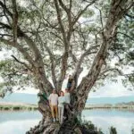 Four people stand on and around a large tree by a lake, with mountains in the background.