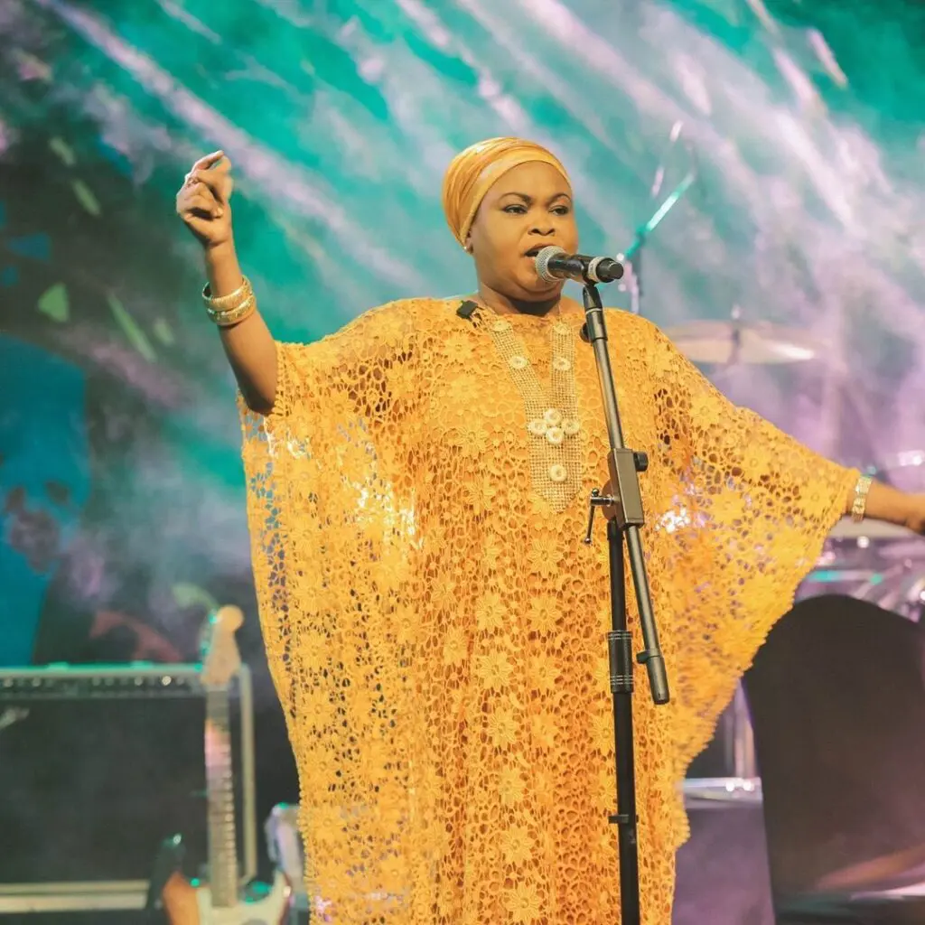 A performer sings on stage, wearing a vibrant orange dress and headscarf. Musical instruments are visible in the background amidst colorful lighting.