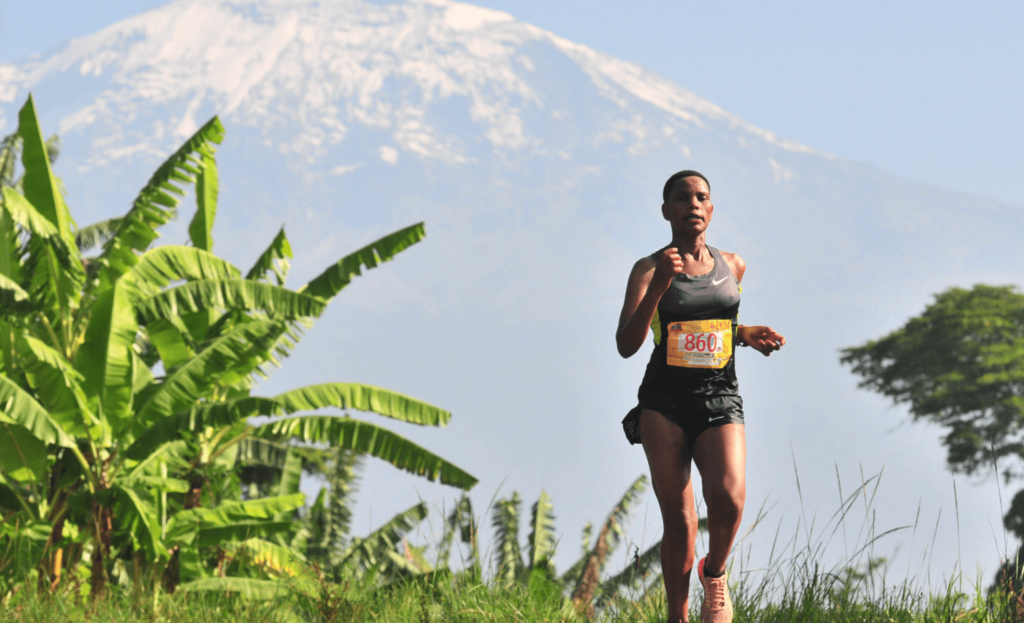 A runner wearing a race bib jogs on a grassy path with a snow-capped mountain and lush green plants in the background.