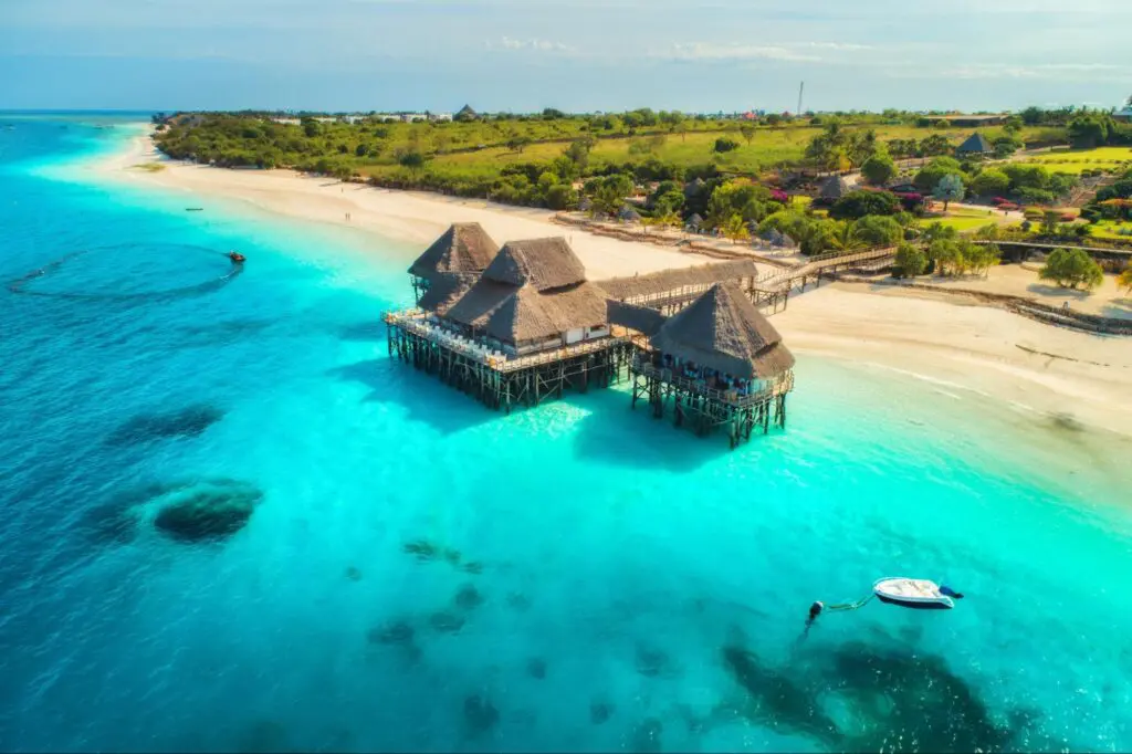 Aerial view of a tropical beach with turquoise water, thatched roof huts on stilts, a small boat nearby, and lush greenery in the background.