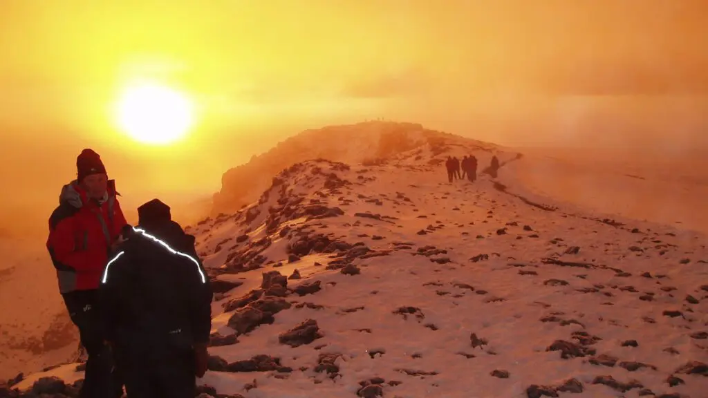 Hikers on a snow-covered ridge during a vibrant orange sunset, with the sun partially obscured by clouds.