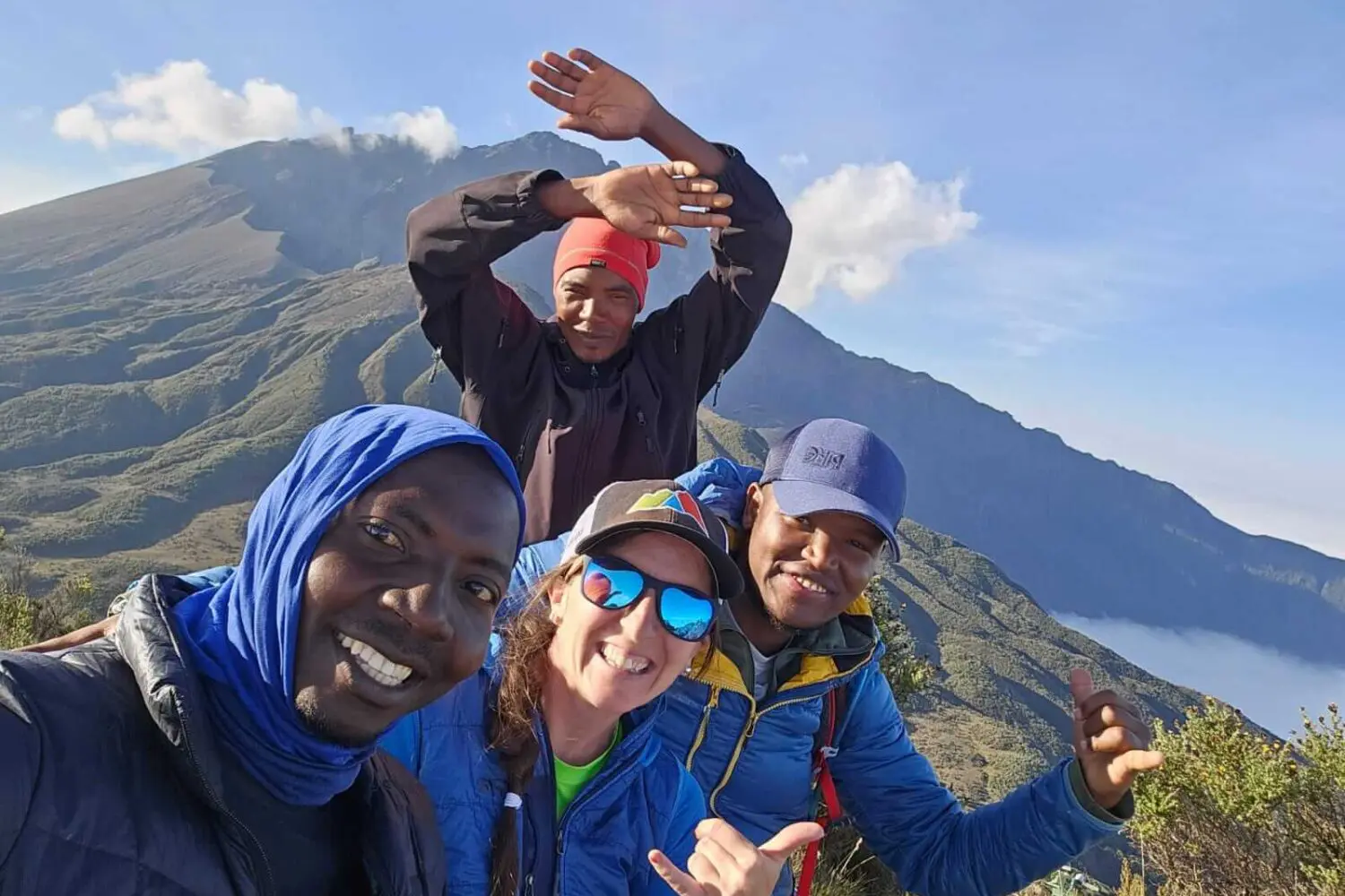 Four people smiling and posing for a selfie atop Mt Meru, capturing the breathtaking landscape during their trekking tour.