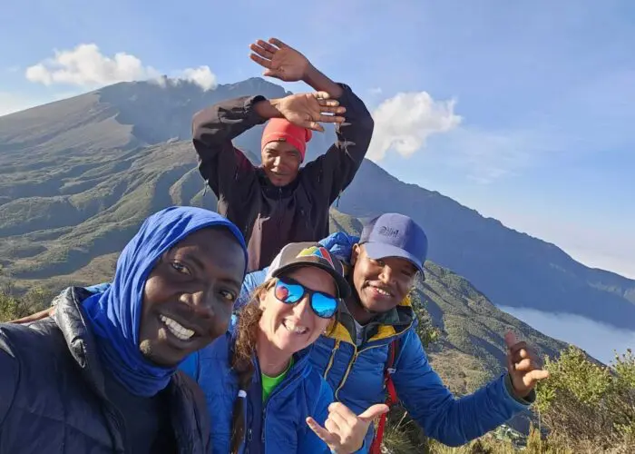 Four people smiling and posing for a selfie atop Mt Meru, capturing the breathtaking landscape during their trekking tour.