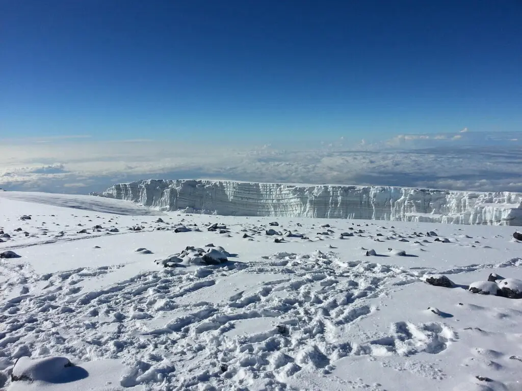 Snow-covered landscape with rocky terrain in the foreground and an ice formation on the horizon under a clear blue sky.