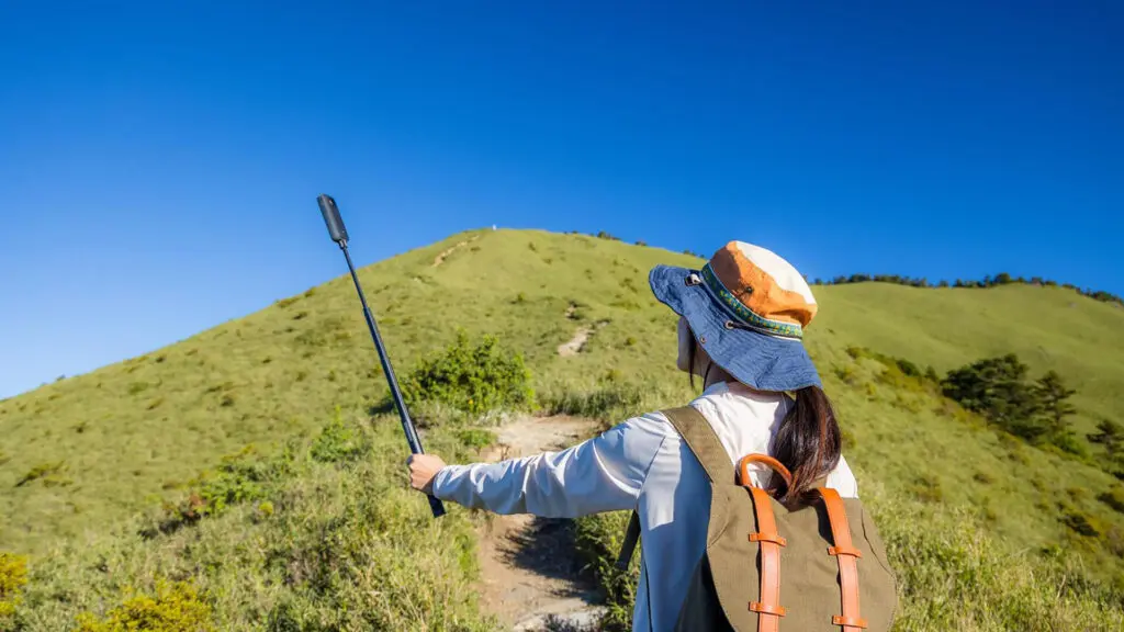 A person wearing a hat and backpack takes a selfie with a selfie stick while standing on a grassy hill under a clear blue sky, capturing their Kilimanjaro training journey for first-timers following a new fitness plan.