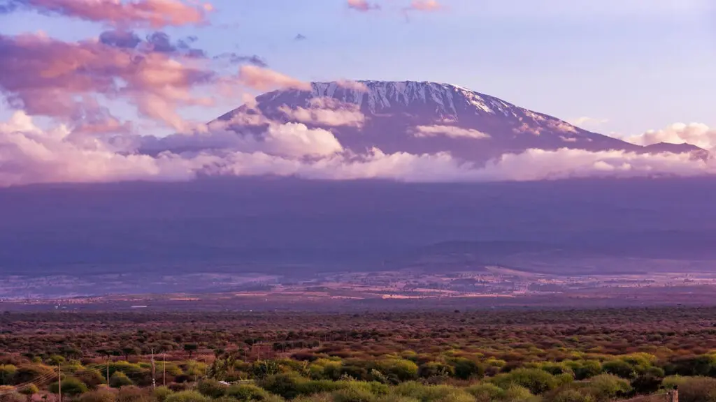 A snow-capped mountain peak rises above a layer of clouds, with a vast plain and sparse vegetation in the foreground—an inspiring scene for first-timers beginning their Kilimanjaro training under a partly cloudy sky.