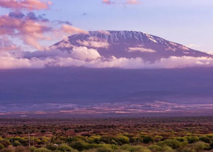 A snow-capped mountain peak rises above a layer of clouds, with a vast plain and sparse vegetation in the foreground—an inspiring scene for first-timers beginning their Kilimanjaro training under a partly cloudy sky.
